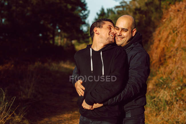Веселая гомосексуальная пара, обнимающаяся в лесу в солнечный день на размытом фоне — стоковое фото