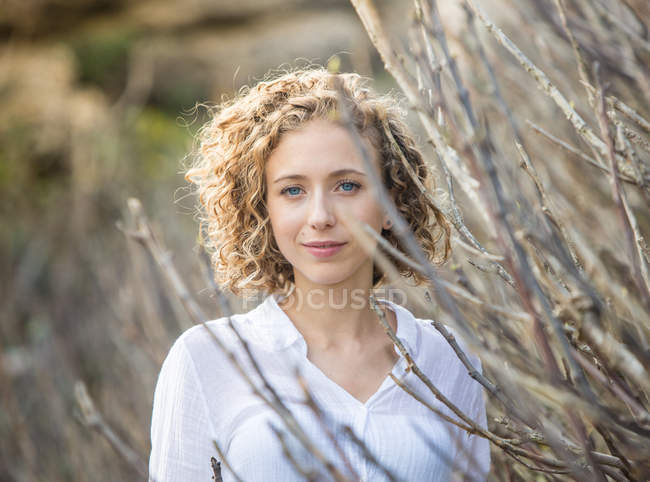 Junge charmante Frau blickt in die Kamera in der Nähe trockener Strauchzweige auf verschwommenem Hintergrund — Stockfoto