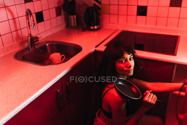Sensual young brunette woman in lingerie looking at camera and holding frying pan in kitchen between redness — Stock Photo