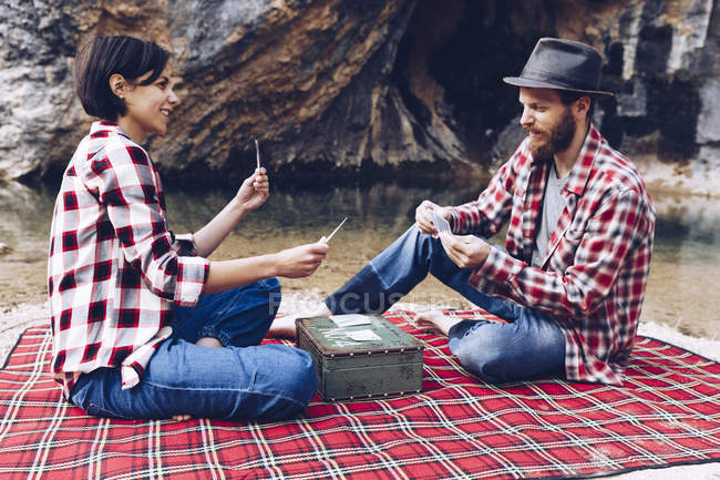 Side view of man and woman in plaid shirts playing cards on plaid having picnic on shore of lake in cliffs — Stock Photo