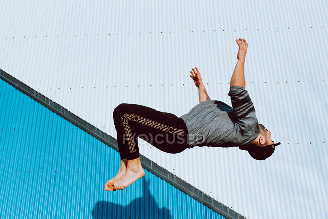 Босоногий парень в стильном наряде, выполняющий сальто возле стены современного здания на городской улице — стоковое фото