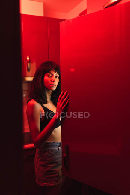 Sensual jovem morena em lingerie olhando para a câmera perto da geladeira na cozinha entre a vermelhidão — Fotografia de Stock