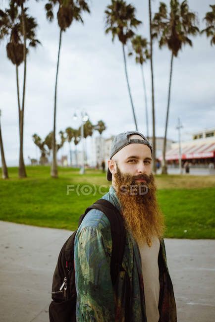 Вид збоку бородатого хлопця з рюкзаком, який дивиться на камеру, стоячи на тротуарі на вулиці сучасного міста — стокове фото