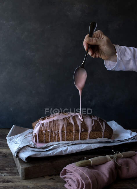 Crop mano di umano con cucchiaio versando topping su torta fresca gustosa arancione posto sul tovagliolo vicino asciugamano e spazzola su sfondo nero — Foto stock