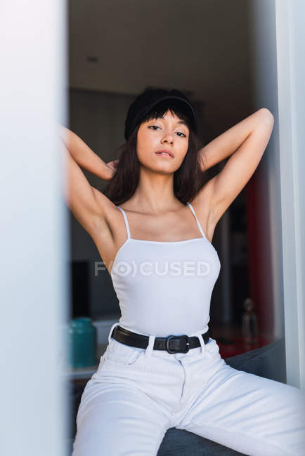 Young slim woman in cap looking at camera and standing near big window — Stock Photo