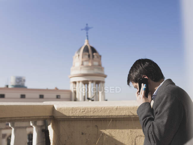 Vista laterale del giovane maschio che parla sul telefono cellulare sulla terrazza vicino alla chiesa nella giornata di sole — Foto stock