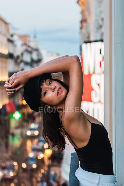 Young slim woman in cap looking at camera and standing on balcony near street with lights in evening — Stock Photo