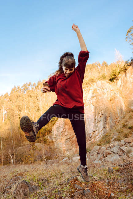 Joven mujer hipster con la mano levantada escuchando música en el teléfono móvil y bailando en la naturaleza - foto de stock
