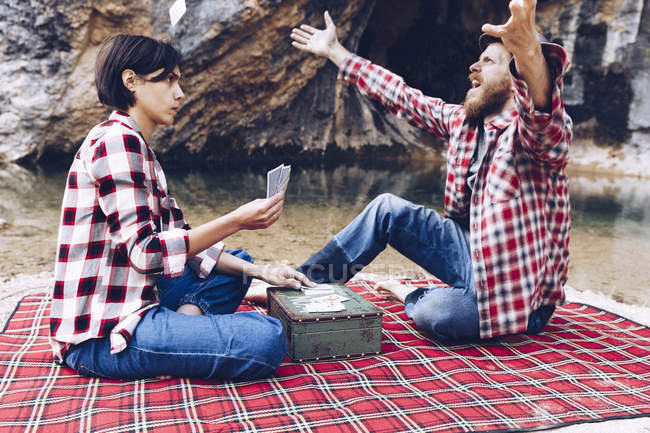Vista laterale di uomo e donna in camicie a quadri giocare a carte sul plaid fare picnic sulla riva del lago in scogliere — Foto stock