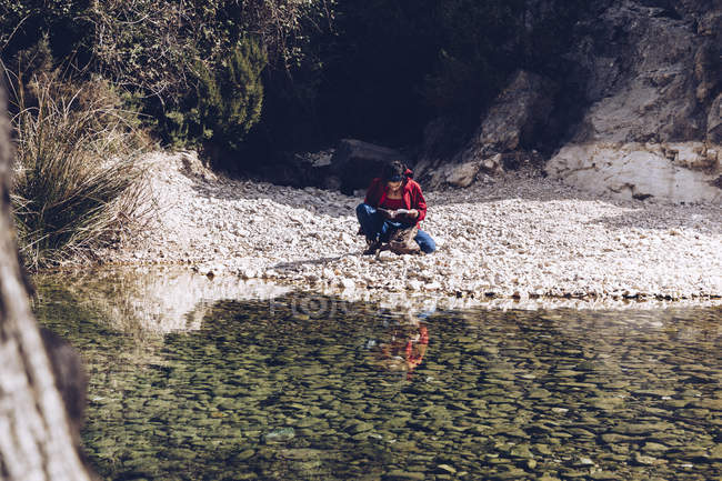 Jovem mulher sentada na costa rochosa do rio de montanha perto de penhasco — Fotografia de Stock