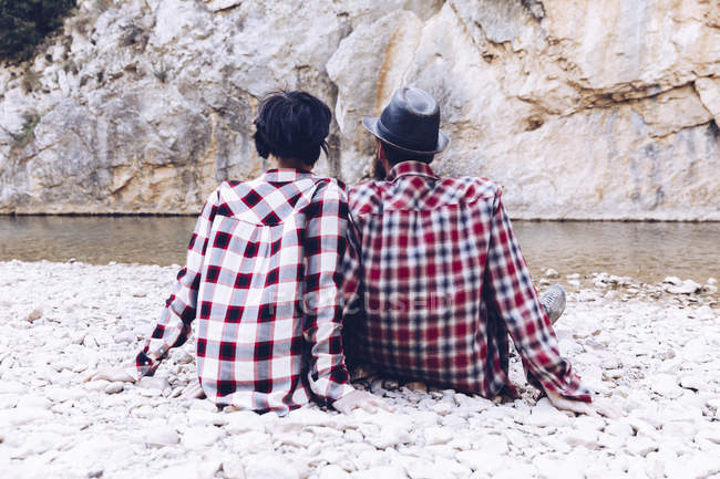 Back view of young couple sitting on rock coast of mountain river near cliff — Stock Photo