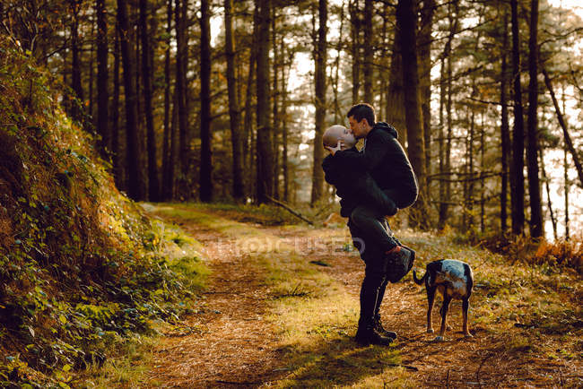 Homosexual couple embracing and kissing near dog on route in forest in sunny day — Stock Photo