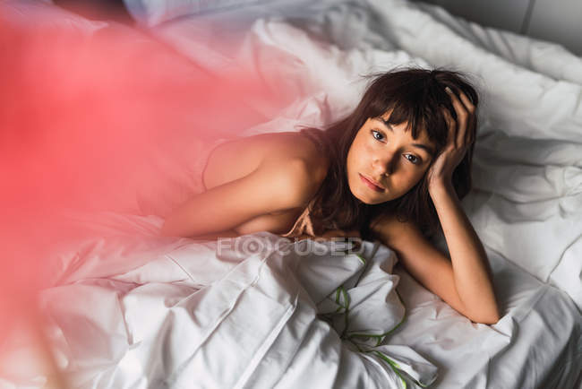 Junge attraktive Frau blickt in die Kamera, während sie sich im Schlafzimmer auf dem Bett ausruht — Stockfoto