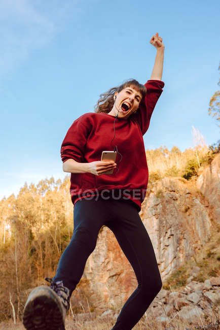 Joven mujer hipster con la mano levantada escuchando música en el teléfono móvil y bailando en la naturaleza - foto de stock