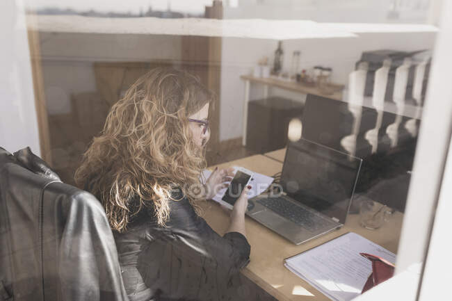 Vue latérale de la jeune femme naviguant sur téléphone portable et assise à table avec ordinateur portable dans le bureau par la fenêtre — Photo de stock