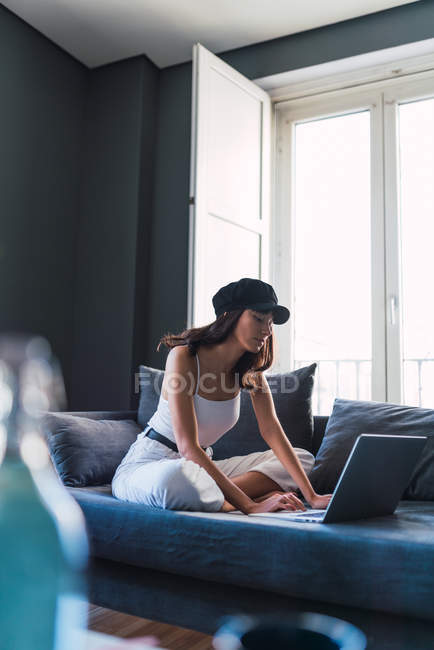 Attractive young stylish woman in cap using laptop while resting on sofa near balcony at home — Stock Photo