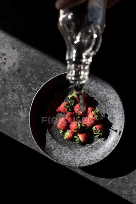 Abgeschnittene Hand einer Person, die eine Flasche hält und Wasser aus frischen Erdbeeren in eine Schüssel auf dunklem Hintergrund gießt — Stockfoto