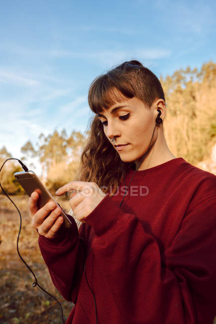 Mulher jovem hipster com piercing e fones de ouvido ouvindo música com telefone celular e andando na estrada rural — Fotografia de Stock