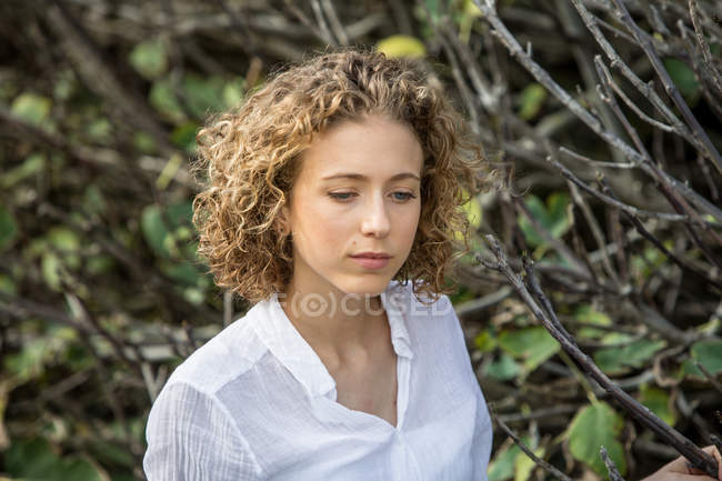 Jeune femme réfléchie posant près de branches sèches d'arbuste sur fond flou — Photo de stock