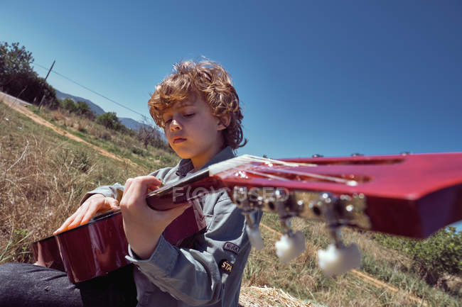 Милый мальчик играет на акустической гитаре и поет, сидя в огромном рулоне сухого сена в солнечный день в сельской местности — стоковое фото