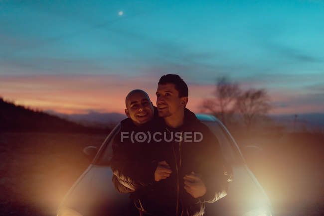 Feliz pareja homosexual abrazándose en el prado cerca del automóvil con faros iluminados en la noche - foto de stock