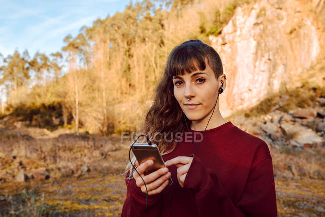 Young woman with piercing and earphones listening music on mobile phone in countryside — Stock Photo