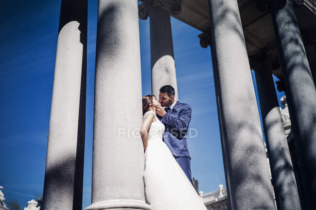 From below side view of young elegant couple in wedding dresses kissing near columns on street in sunny day — Stock Photo