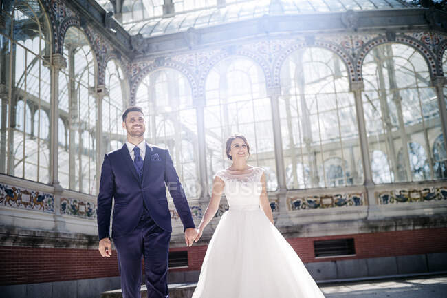 Von unten hält ein junger eleganter Mann eine Frau im Hochzeitskleid in der Nähe des Retro-Palastes mit vielen Fenstern an sonnigen Tagen an den Händen — Stockfoto