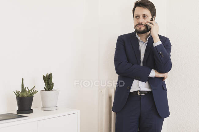 Hombre joven concentrado con la mano cruzada hablando por teléfono móvil en la habitación con planta de interior y libro en la mesa - foto de stock