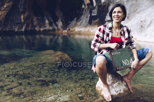Woman sitting on rock in transparent lake water looking inside of rusty old metal case having picnic — Stock Photo