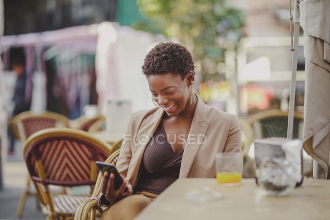 Cheerful African American elegant woman holding mobile phone and sitting at table with glass of juice near baggage in street cafe — Stock Photo