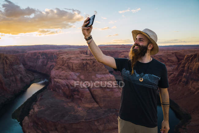 Hombre barbudo guapo sonriendo y tomando selfie mientras está parado contra magnífico cañón y río durante el atardecer en la costa oeste de EE.UU. - foto de stock