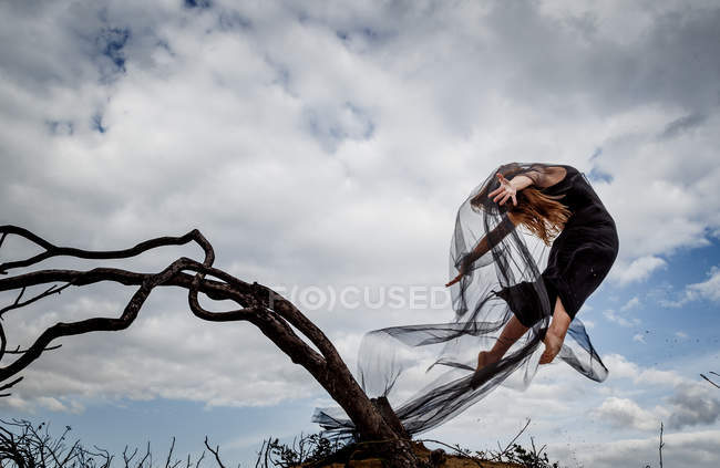 De baixo da bailarina jovem no desgaste preto com as mãos levantadas no ar perto de ramos secos e céu azul em nuvens — Fotografia de Stock