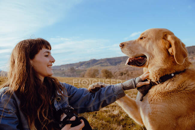 Vista lateral de mujer joven disparando en cámara perro divertido entre prado y cielo azul - foto de stock