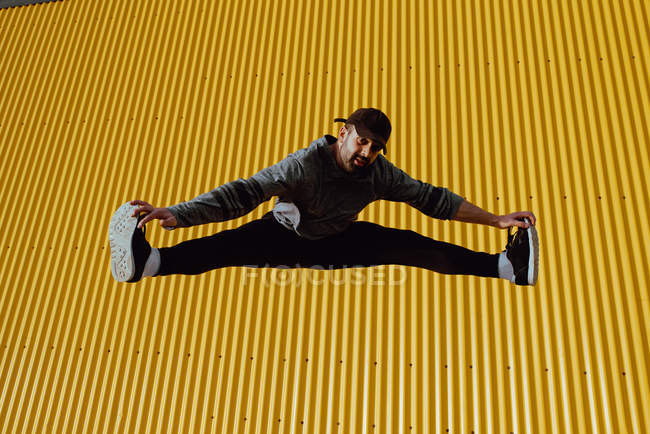 Бородатый парень в модном наряде прыгает и смотрит в камеру на желтую стену современного здания в солнечный день — стоковое фото