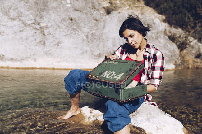Woman sitting on rock in transparent lake water looking inside of rusty old metal case having picnic — Stock Photo