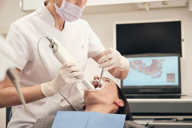 Mujer en guantes y mascarilla utilizando equipos modernos para hacer escaneo de dientes de paciente femenina en el consultorio del dentista - foto de stock