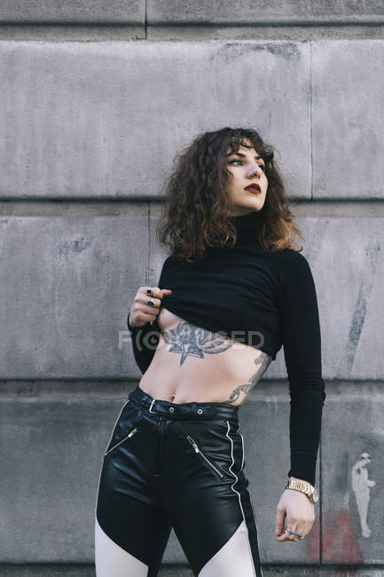 Attractive young woman lifting stylish sweater and showing tattoo while standing near building wall on city street and looking away — Stock Photo