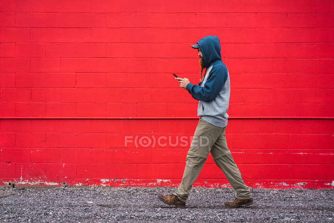 Вид збоку хлопця з плетеною бородою на смартфоні для перегляду пуховиків під час ходьби біля червоної стіни на міській вулиці — стокове фото