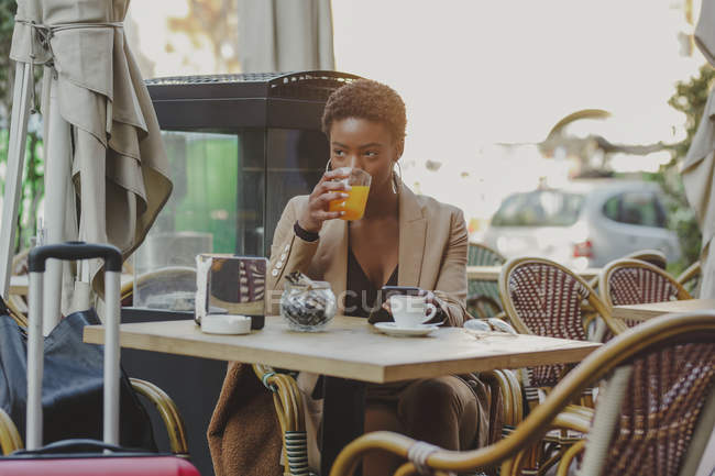 Afro-américaine élégante femme tenant téléphone portable et boire du jus près des bagages dans un café de rue — Photo de stock