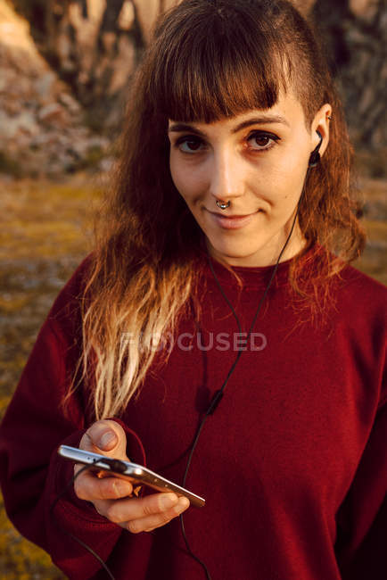 Jovem mulher hipster sorridente com piercing e fones de ouvido ouvindo música com telefone celular no campo — Fotografia de Stock
