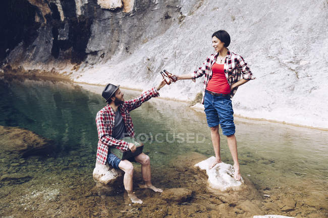 Man and woman in transparent water of beautiful lake on rocks clinking with beer bottles enjoying picnic — Stock Photo