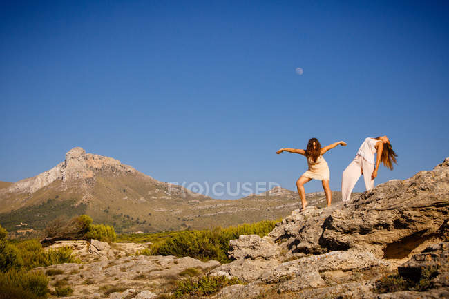 Mujeres jóvenes misteriosas con las manos levantadas posando en rocas cerca de la colina y el cielo azul con la luna - foto de stock