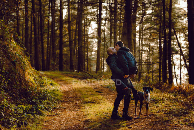 Гомосексуальная пара обнимается и целуется рядом с собакой по пути в лес в солнечный день — стоковое фото