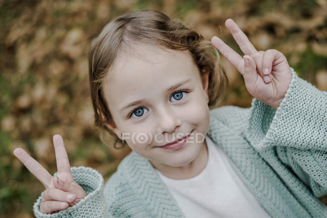 Позитивная маленькая девочка смотрит в камеру в парке с сухой листвы и показывает признаки победы — стоковое фото