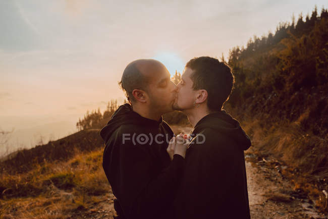 Happy homosexual couple embracing and kissing on route in forest in sunny day — Stock Photo