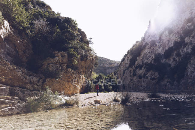 Вид ззаду жінка з рюкзаком на узбережжі гірської річки біля скельної гори — стокове фото