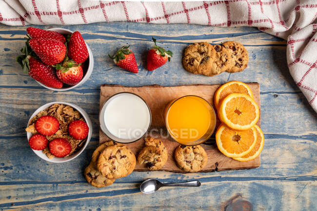 From above glasses of milk and juice near spoon, tasty biscuits, slices of orange and strawberries on wooden background — Stock Photo
