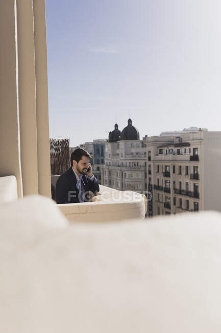 Молодий чоловік розмовляє по мобільному телефону на балконі біля споруд міста в сонячний день. — стокове фото