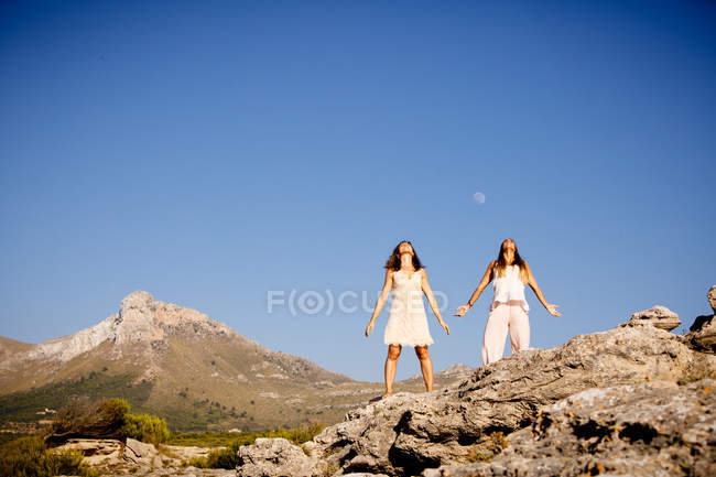 Jovens senhoras misteriosas posando em rochas perto de colina e céu azul com lua — Fotografia de Stock
